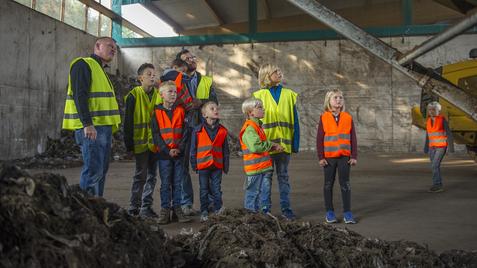 Gruppe von Erwachsenen und Kindern in Sicherheitswesten beobachtet eine Maschine in einer Halle mit Abfallablagerungen.