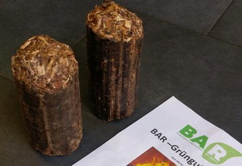 Zwei Grünbriquettes aus Biomasse neben einem Informationsblatt mit Heizwertangaben auf einem grauen Untergrund.