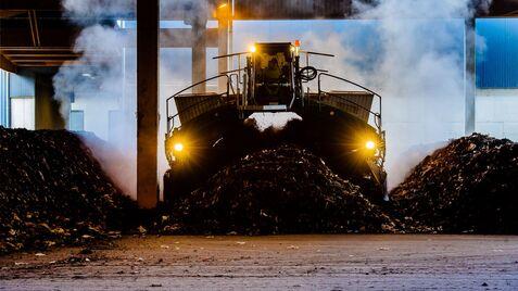 Blick auf einen großen Bagger, der Abfall in einer Halle bewegt, umgeben von Dampf und Müllhaufen.