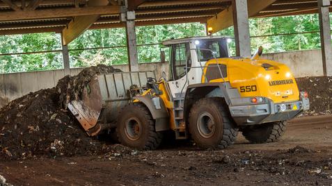 Radlader der Marke Liebherr 550 schaufelt Abfall in einer überdachten Halle mit grüner Vegetation im Hintergrund.