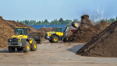 Baufahrzeuge arbeiten auf einem Gelände mit großen Komposthaufen, im Hintergrund Windräder und Solarpanels sichtbar.