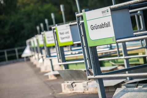 Reihe von Abfallstationen mit Beschilderung für Grünabfall und Restmüll in einem Recyclingbereich.