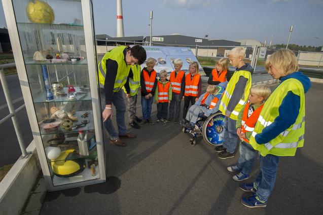 Ein Mann deutet mit der Hand auf Dinge in einem Glasschaukasten. Eine Gruppe von Kindern in gelben und orangen  Westen sieht interessiert zu.