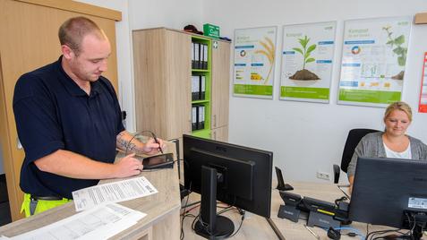 Mann am Empfang schreibt auf ein Tablet, während eine Frau am Computer arbeitet. Im Hintergrund Grafiken zu Kompost.