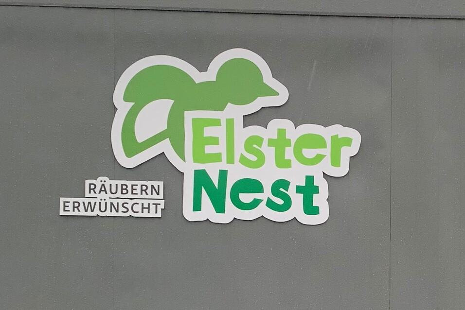 Schild mit der Aufschrift "Elsternest" und "Räubern erwünscht" an einer grauen Wand, im Hintergrund ein Windrad.