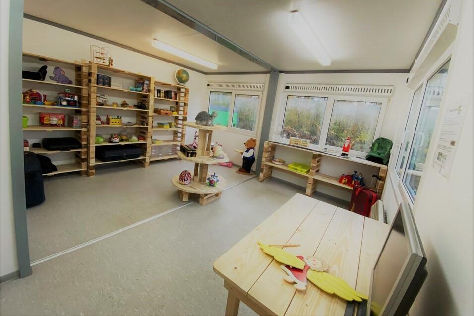 Innenansicht eines Spielraums mit Regalen, Spielzeug, einem Tisch und Fenstern, die auf einen grünen Außenbereich blicken.