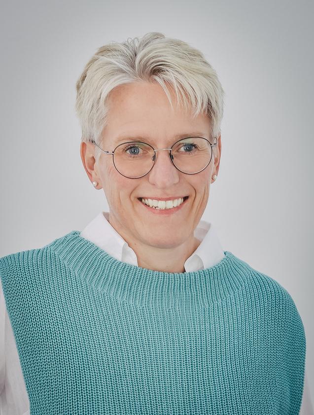 Lächelnde Frau mit kurzen, blondierten Haaren, Brille und türkisfarbenem Pulli vor neutralem Hintergrund.