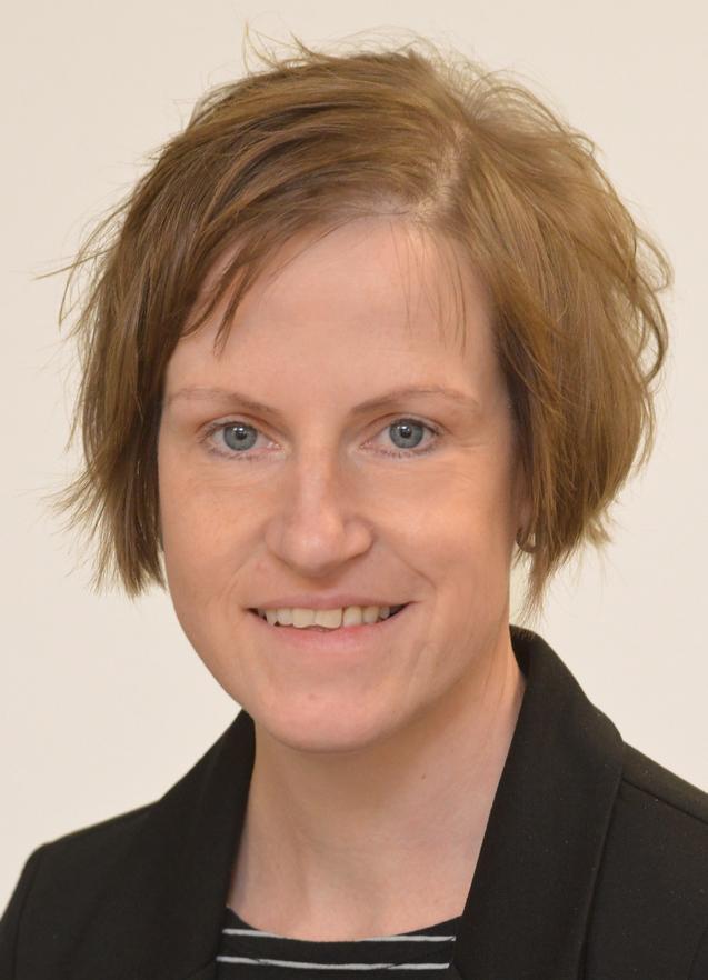 Porträt einer Person mit kurzen, braunen Haaren, die lächelt und einen schwarzen Blazer sowie ein gestreiftes Oberteil trägt.