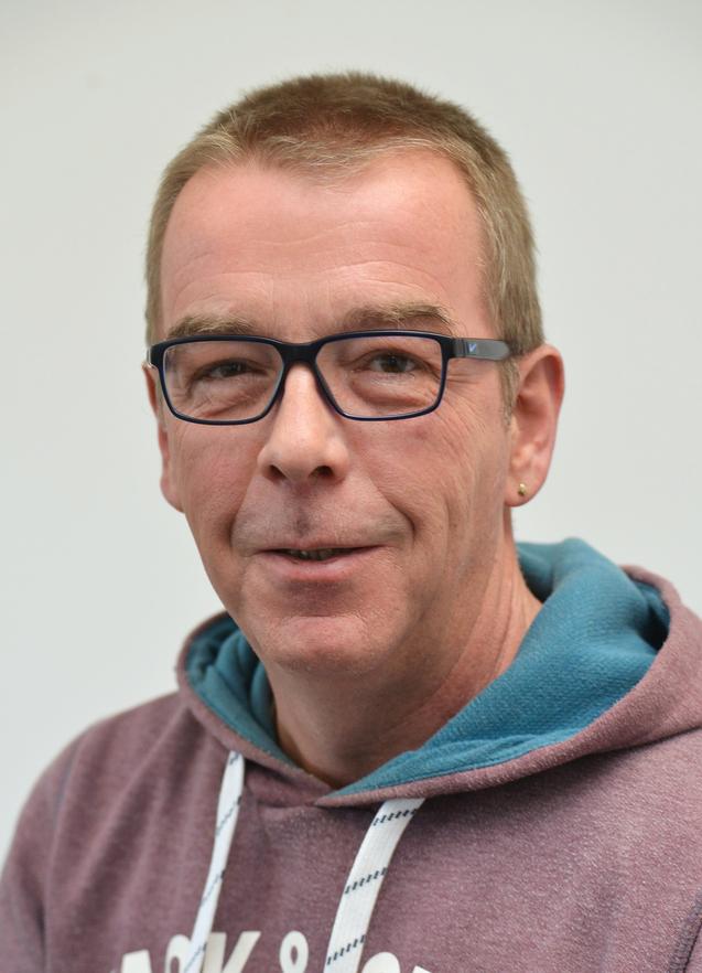 Mann mit Brille, kurzen blonden Haaren und einem schlichtem Kapuzenpullover lächelt vor einem hellen Hintergrund.